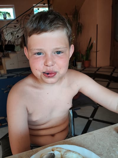 A young boy is shown from the chest up, looking directly at the camera. He has short, brown hair and blue eyes. His lips are stained purple, suggesting he has recently eaten something like berries or candy. He is shirtless, and his skin is fair with some freckles. In the background, a staircase with an ornate metal railing is visible, leading up to two windows. There are potted plants in the background, and the floor has a tiled pattern. A table with a marbled surface is in the foreground, with a plate of food and a spoon on it. The lighting suggests it is daytime, likely indoors. The overall setting appears to be a home in a residential area.