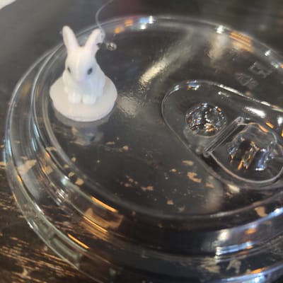 A small white ceramic bunny figurine sits on a white circular base, which is placed on a clear plastic lid. The lid is resting on a dark, textured surface that appears to be a table. The bunny is positioned on the left side of the lid, facing slightly to the right. Its ears are upright, and its eyes are dark. The lid has raised ridges and a molded drinking spout area on the right side. Light reflects off the lid and the bunny, creating highlights and some glare. The dark surface beneath the lid has a distressed appearance with lighter speckles. The overall impression is a close-up shot of a decorative item on a container lid. There is no information about Kobeliaky, Ukraine in the image.