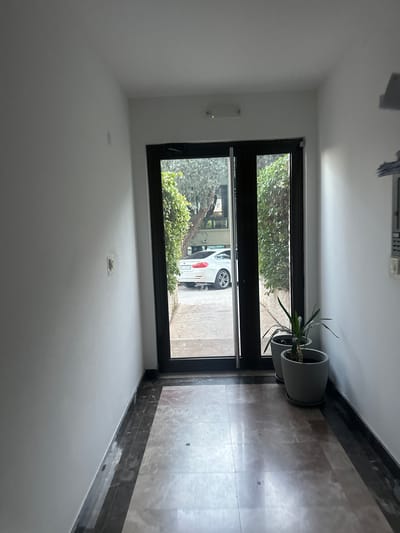 The image depicts an interior view of a hallway or entryway in Podličak, Montenegro, looking out through a large glass door or window. The walls are painted white, and the floor is covered with polished, light brown or beige marble-patterned tiles, bordered by a darker trim.
Through the black-framed glass, green foliage from outdoor bushes or trees is visible. A white car, possibly an SUV or hatchback, is parked on a paved surface directly outside. The car's license plate clearly displays the text "E4 0303".
To the right of the glass door/window, two grey cylindrical planters, each holding a green potted plant, are placed on the tiled floor. On the white wall to the left, a slender, vertical light fixture is mounted, and below it, a small square sensor or indicator is affixed to the wall. Above, a ceiling fixture, possibly an air vent or light fitting, is partially visible. The scene is brightly lit, indicating daytime. No human activity is present.