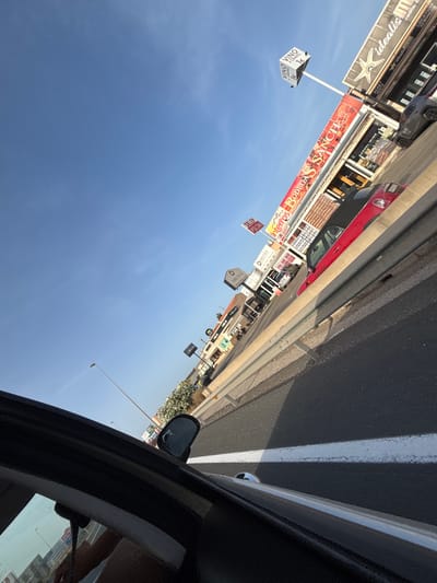 The image shows a road scene in Urbanización Cabo Roig, Spain, with a bright blue sky overhead. The perspective is from inside a vehicle, looking outwards. A metal guardrail runs along the side of the road. Beyond the guardrail, there are several shops and businesses with visible signage. One sign reads "Bodegas SANCH", another has "VINO A CHANCE 1€" on a cube-shaped structure. There are red and grey cars parked or driving along the road. The buildings appear to be low-rise commercial structures. In the foreground, the interior of the vehicle is visible, including a side mirror and part of the dashboard. The lighting suggests it is daytime, likely late afternoon due to the angle of the shadows.