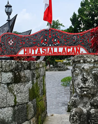 The image depicts the entrance to Hutasiallagan in Ambarita, Indonesia. A decorative red and black archway with white lettering spelling "HUTASIALLAGAN" spans the foreground. To the left, a stone wall covered in moss stands beside a lamppost and a portion of a traditional roof. To the right, a weathered stone statue with a human-like face is partially visible, also showing signs of moss growth. Beyond the archway, a paved courtyard is visible, leading further into the site, with trees and foliage in the background. A red and white flag is prominently displayed above the archway. The sky is overcast with clouds.
