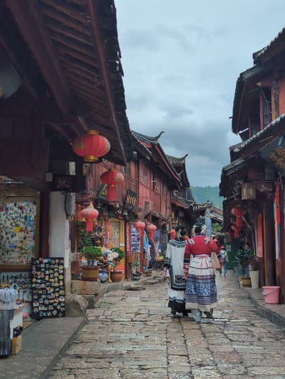 The image depicts a street scene in a traditional Chinese town, likely Lijiang, given the architectural style. The perspective is vertical, as if the camera were held sideways. Red, multi-story buildings with traditional tiled roofs line a narrow, cobblestone street. Red lanterns are suspended from the eaves of the buildings, adding a festive and cultural element.
Several people are visible, dressed in what appears to be traditional ethnic attire. One individual in the foreground wears a red top and a patterned skirt. Another person beside them wears a white top with intricate black and white patterns. They are walking along the street, suggesting movement and daily life.
The street is lined with shops showcasing various goods. Shelves display an array of items, including what look like souvenirs or local crafts such as magnets and figurines. The shops appear to be open, with merchandise displayed outside.
The weather seems overcast, with a cloudy sky dominating the upper portion of the image. The lighting is diffused, suggesting it might be late afternoon or early morning, or simply a cloudy day. The overall atmosphere is one of a bustling historic marketplace, with a strong sense of local culture and tradition. No discernable text is legible in the image.