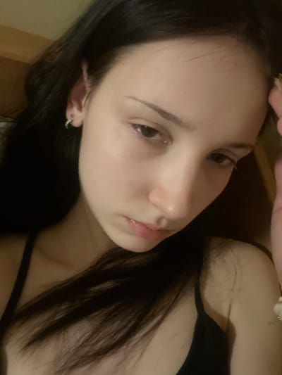 A close-up, angled portrait features a young woman with fair skin, dark eyes, and long, dark hair, which frames her face. Her head is tilted, and she looks towards the camera with a neutral, slightly pensive or contemplative expression, her lips gently parted. She wears a dark-colored top with a strap visible over her right shoulder. A small silver hoop earring is visible in her left ear. At the top of the frame, a hand, possibly her own, rests lightly on her forehead or hair, showing a glimpse of a light-colored bracelet or accessory. The lighting is soft and warm, suggesting an indoor setting, possibly in dim or evening light. The background is out of focus, indicating a shallow depth of field. There is no visible text in the image.