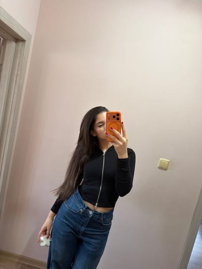 The image displays an indoor mirror selfie of a young woman, captured with the photo rotated 90 degrees clockwise.

**Subjects:** The central subject is a woman with long, straight, dark brown hair. She is wearing a fitted black long-sleeved top with a silver zipper down the front and blue denim jeans. Her fingernails are painted a vibrant red. She is holding an orange smartphone in her right hand, which features a circular MagSafe accessory on its back. In her left hand, she holds a white, flower-shaped hair clip.

**Activity or event taking place:** The woman is actively taking a self-portrait using a mirror, holding her phone to capture her reflection. Her gaze is directed towards the phone.

**Notable details:** The background is a plain, light beige or pale pink wall. A classic-style light switch with a yellowish plastic cover is visible on the wall to the upper right. Part of a doorway with light-colored wooden trim is seen in the lower left corner, and a glimpse of wooden flooring appears in the lower right. The woman's attire is casual, and the room appears to be a simple home interior. The bright orange phone case and red nail polish provide a splash of color against her darker clothing. There are no clear cues for the time of day or specific weather conditions. The scene suggests an everyday, relaxed moment.

**Visible text:** No visible text is present in the image.