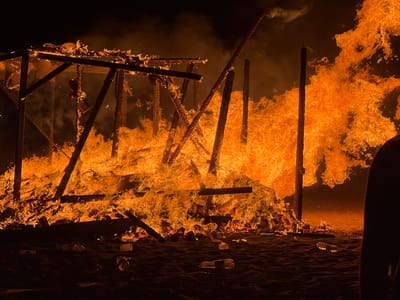 An open-air structure, possibly a temporary construction, is engulfed in intense flames. The fire is consuming the wooden beams and supports, creating a dynamic and chaotic scene with a significant amount of smoke and heat. The flames are primarily orange and yellow, with some white areas indicating extreme heat. The ground appears to be sandy, with several plastic bottles and cups scattered across it, suggesting a public event or gathering. The background is dark, indicating nighttime. The overall impression is one of destruction and intense heat. The image appears to have been taken in the city of La Línea de la Concepción, Spain.