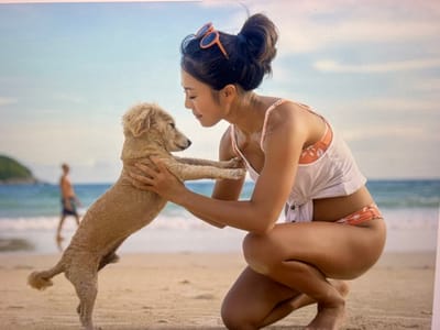 A young Asian woman with dark hair pulled up in a messy bun and orange sunglasses perched on top is kneeling on a sandy beach. She is wearing a white tank top over a patterned bikini top and matching bikini bottoms. She is holding a small, light-colored dog, which is standing on its hind legs and looking at her. The woman is looking down at the dog with a gentle expression. In the background, the ocean meets the shore, with waves gently lapping the beach. Further out, a blurred figure of a person in swim trunks can be seen walking along the waterline. The sky above is a pale blue with wispy clouds. The image does not depict the city of Bevern, Germany.