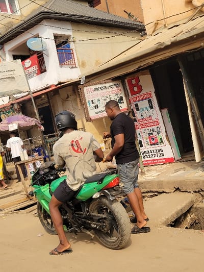 This image depicts an outdoor street scene in Puerto López, Colombia, under daylight conditions. Two adult males are central to the foreground. One male, wearing a black helmet, light grey t-shirt with a red and black logo on the back, black shorts, and black flip-flops, is seated on a bright green sport motorcycle, facing away from the viewer. The motorcycle features black wheels and a black seat. Adjacent to the motorcycle, a second male stands to its right, wearing a black t-shirt, blue denim shorts, and black slip-on shoes. He holds a yellow object in his right hand, facing towards the motorcycle.
The immediate ground consists of uneven dirt and concrete, with a notable break in a concrete slab exposing a dark void on the right. In the background, multiple commercial buildings line the street. The building directly behind the standing man features multiple signs, including one with red and black text advertising "FLASHING," "UNLOCKING," and "ACCESSORIES" for mobile devices, along with phone numbers "00341286" and "03222726." Another building further left is two-storied, featuring a white upper facade with a blue satellite dish and a window with a blue railing. A red sign reading "NEWELL" is visible on the lower section of this building. A utility pole with overhead wires is visible on the far left. Further into the background on the left, additional individuals and a purple-and-white striped umbrella are partially visible.