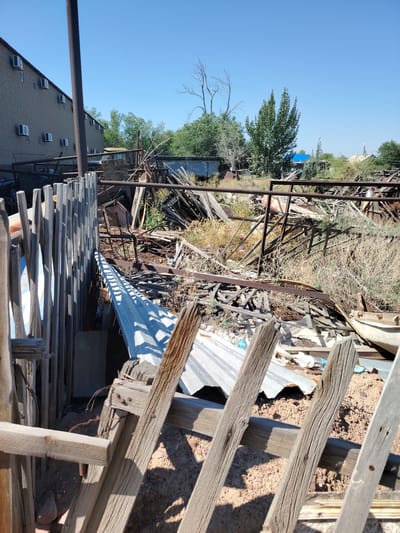 The image depicts a cluttered outdoor area, likely a junkyard or storage yard, in the vicinity of Тораңғылық, Kazakhstan. In the foreground, a weathered wooden fence constructed from vertical planks casts sharp shadows. Behind the fence, a chaotic jumble of scrap metal, including corrugated roofing sheets and various structural components, is piled high. Dry brush and weeds are interspersed throughout the debris. To the left, a portion of a large, multi-story brick building with numerous air conditioning units mounted on its facade is visible, suggesting an urban or semi-urban setting. The sky above is clear and blue. The scene is devoid of any people or animals. The overall impression is one of disarray and abandonment.