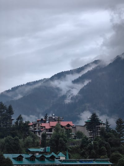 An overcast, cloudy day looms over the hilly terrain of Aleo, India. Dense, dark green pine trees blanket the slopes of a large mountain that dominates the background. Wisps of white fog weave through the trees, partially obscuring the mountain's contours and creating a dramatic, ethereal effect. In the midground, several multi-story buildings with red-tiled roofs are nestled amongst the trees. These structures appear to be hotels or guesthouses, indicative of a tourist destination. One prominent building features a gray facade with visible balconies. Further down in the foreground, more buildings with green, pitched roofs are visible, suggesting a lower-lying residential or commercial area. The overall atmosphere is one of subdued natural beauty, with the heavy clouds and mist contributing to a sense of quiet and isolation. No people or animals are visible in the frame, and no specific actions or interactions are taking place. The scene is static and focused on the landscape and architecture.