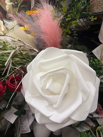 This is a close-up, eye-level, artificial light, medium shot of a very large white artificial rose with thick, curved petals. The rose is the dominant subject, filling most of the frame from the bottom left to the top right. To the left of the rose, there are two smaller red artificial roses with visible stems and leaves. Behind and to the left of the main rose, a variety of artificial floral elements are visible, including pink feathery plumes, white feathery plumes, green grass-like elements, and twigs with white beaded accents. In the background, to the right of the rose, there are shelves with hanging green artificial plants and woven baskets. Several product tags with barcodes and text are attached to the stems of the artificial flowers. There are no people visible in the image. The scene appears to be a retail display, likely in a store that sells artificial flowers and home decor. There is no indication of the image being taken in Würzburg, Germany, or any specific city.