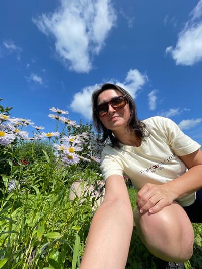 A woman is taking a selfie in a garden with flowers under a blue sky with white clouds. She is wearing sunglasses and a cream-colored t-shirt with a Tommy Hilfiger logo. Her right hand is resting on her knee. The garden is full of green foliage and light purple flowers. The location is identified as Kumyony, Russia.