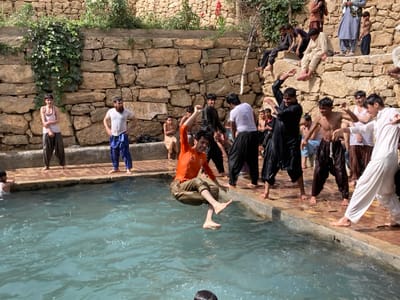The image depicts an outdoor recreational scene in Killi Malik Sher Mohammad, Pakistan, featuring approximately 20-25 males of varying ages, from adolescents to adults, gathered around a rectangular-shaped pool of light blue-green water.
In the foreground and mid-ground, a young male wearing an orange long-sleeved shirt and light-colored trousers is captured mid-air, actively jumping into the pool, his bare feet elevated above the water's surface, creating splashes. To the immediate right of the pool, several other males are engaged in dynamic activities on the paved edge. One male in a wet, black shalwar kameez has both arms raised in a dancing or celebratory pose. Adjacent to him, a bare-chested male in dark, wet trousers displays a similar energetic stance. Further to the right, a male in a white shalwar kameez also participates in the lively interaction. Other figures, some bare-chested and some in wet traditional attire, are positioned along the pool's edge.
On the left side of the pool, a bearded male, wearing a wet white t-shirt and blue patterned trousers, stands with hands on his hips, observing the water. To his left, a younger male in a light-colored tank top and dark trousers stands still. A partial view of another individual submerged in the water is visible on the far left.
The background is dominated by a multi-tiered stone wall, constructed from irregular, light-colored stones, rising behind the pool. Green vegetation, including climbing vines and some red flowering plants, grows on and above the stone structure. Multiple males are seated or standing on various ledges of this stone wall, observing the activities below. They are dressed in traditional Pakistani attire, including shalwar kameez in beige, light blue, and black, with some individuals bare-chested. The ground immediately surrounding the pool is paved with light brown or reddish tiles. The scene conveys a communal gathering centered on water-based recreation.