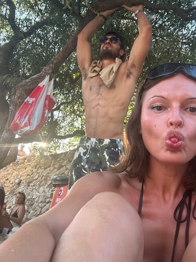 The image depicts an outdoor leisure scene, likely a shaded rocky beach or recreational area in Tisno, Croatia, during daylight hours.

In the foreground, a female with medium-length brown hair faces the camera in a close-up selfie. She has dark sunglasses pushed onto her forehead, and her tanned skin appears wet. Her mouth is puckered in a "kissing" pose, with the tip of her tongue slightly visible. She wears a black string bikini top, and her right shoulder and knees are visible, indicating a seated or reclined position.

Behind her, in the mid-ground, a shirtless male with dark hair, a beard, sunglasses, and a backward cap is positioned. He displays a muscular physique with visible abdominal muscles and body hair. He wears camouflage-patterned swim shorts with a white drawstring, and a light-colored cloth is draped around his neck. His arms are raised, gripping a thick, gnarled tree branch above his head, suggesting he is hanging or stretching. Beaded bracelets are visible on his left wrist.

The setting is beneath the dense, green canopy of a large tree. To the left of the male, a white plastic bag with a red maple leaf design (resembling a Canadian flag) hangs from a lower tree branch. The background features a rocky hillside or embankment. A red recycling bin, marked "PET" and displaying a recycling symbol, rests on the rocks behind the foreground subjects. Further back on the rocky terrain, a person with light-colored headwear is partially visible, illuminated by a strong sun flare. Another female, wearing a black bikini top and sunglasses, is seated to the left on the rocks.