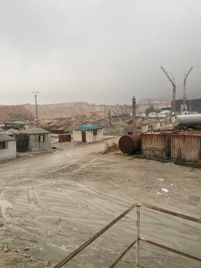 This image captures a desolate industrial or construction site in Dahiyat Harasta, Syria, under an overcast, light grey sky. The foreground features a rusty metal railing extending from the lower right. The ground throughout the scene consists of light brown dirt, extensively marked with tire tracks. In the midground, multiple small, single-story concrete structures are visible; some appear unfinished, while others, like one with a prominent turquoise roof and a brown door, are more complete. Adjacent to these structures are several rusted shipping containers, some stacked, and a large, horizontally-oriented, cylindrical rusted metal tank. Scattered debris, including pieces of paper, is visible on the ground. The background is characterized by extensive excavation or quarrying on hillsides, revealing lighter rock faces. Industrial infrastructure includes two prominent tower cranes, multiple power pylons supporting electrical lines, and distant industrial buildings. No individuals are present, and no active operations or interactions are observable.