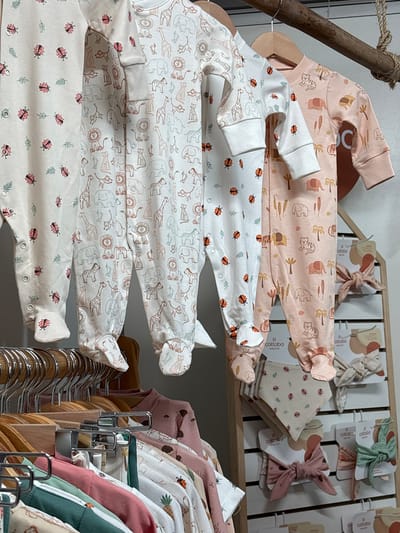 This image shows a display of baby clothing in a retail setting. The clothing consists of baby onesies with various patterns, including animals and ladybugs. Some onesies are stacked on wooden hangers, while others are hanging on a rack. Additionally, there are baby headbands with tags on a shelf in the background. The location appears to be a store specializing in baby products. The image does not provide specific details to confirm the city as Chorrillos, Peru.