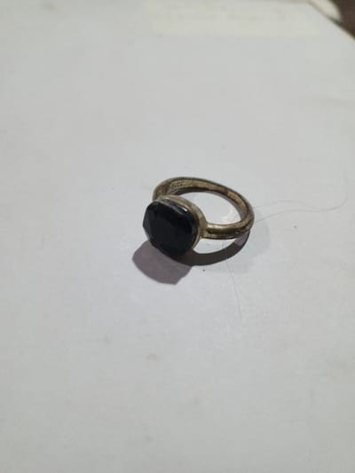 The scene, specified to be within the city of Phakeo, Laos, depicts a single metal ring resting on a uniform, light-colored, flat surface. The ring features a tarnished or oxidized metallic band with a visible texture and an oval-to-octagonal shaped, black, faceted stone set centrally within a bezel. The ring is oriented with its band slightly elevated, casting a defined shadow to its left, indicating a light source from the upper right. Several minute, light-colored fibers, consistent with lint or hair, are visible on the surface near and around the ring. No other subjects, actions, interactions, or specific environmental details beyond the immediate surface are discernible to corroborate the specified location.