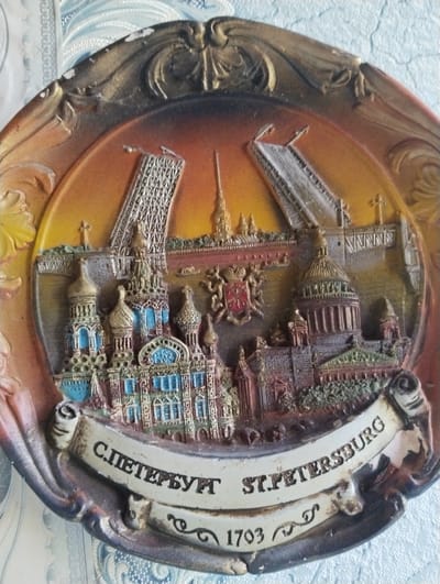 The image shows a decorative plate depicting famous landmarks of St. Petersburg, Russia. The plate is circular and has a raised relief design. In the center, iconic buildings such as St. Isaac's Cathedral and Kazan Cathedral are visible, along with what appears to be a representation of the Peter and Paul Fortress or a similar spire. Two large arched structures, possibly depicting bridges, are prominently featured in the background. The plate has a decorative border with ornate carvings. A banner across the bottom of the plate bears the inscription "С. ПЕТЕРБУРГ ST. PETERSBURG 1703". The background behind the plate is a textured light blue and white patterned fabric. There are no people or animals present in the image. The scene is a static representation of architectural landmarks, not a depiction of an actual location in Заполье, Russia, as Заполье is a village, not a city known for these landmarks.