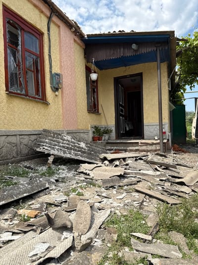 The image displays the exterior of a damaged single-story residential building in Nedryhailiv, Ukraine, under a partly cloudy sky. The building features a yellow stucco facade with a vertical peach-colored textured section on the left and a grey, textured foundation. The ground in front of the building is covered with a large quantity of fragmented, grey, corrugated roofing material, consistent with asbestos cement sheets, intermingled with white fibrous debris and sparse green vegetation. A large, broken sheet of this material leans against the foundation on the left.

The building's two visible windows, framed in red, exhibit shattered glass panes, with shards still present in the left-most window. A dark-colored front door stands open beneath a covered porch that features a dark blue soffit and a decorative wooden valance. A white hanging planter is suspended from the porch ceiling. A grey-green electrical meter box with black conduit is mounted on the yellow wall between the left window and the peach section. Several potted plants are visible near the porch entrance. In the background, green foliage, a partial green gate, and the front section of a white vehicle are discernible on the right. No individuals are present in the scene, and no actions are depicted.