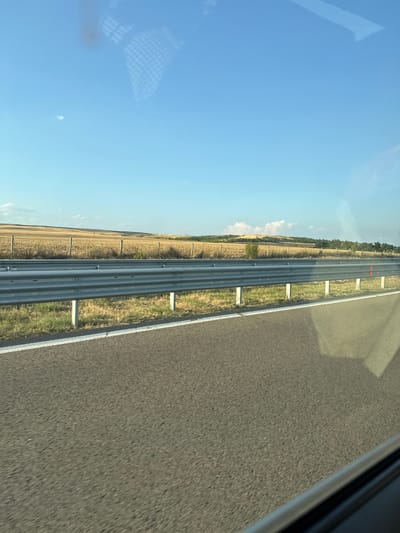 The image depicts a roadside scene with a metal guardrail separating a paved road from a field. The road surface is asphalt, with a white line marking its edge. Beyond the guardrail, a dry, golden field stretches towards rolling hills in the background. The sky is a clear, bright blue with a few wispy white clouds. The scene appears to be from a moving vehicle, as indicated by the slight motion blur on the road surface and reflections on the presumed window. The location is identified as Troyanovo, Bulgaria. There are no people or specific actions visible in the image. The environment is rural and appears to be a sunny day.