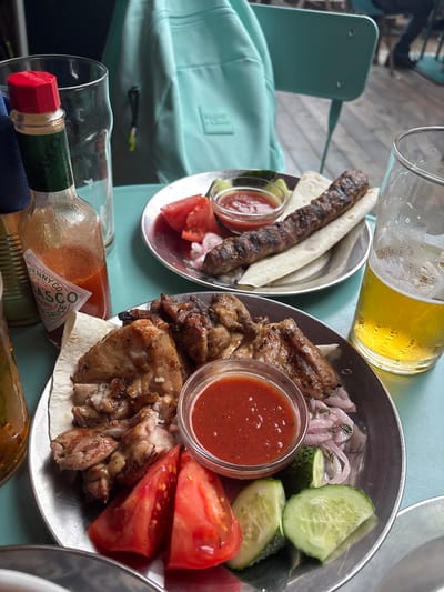 The image depicts a close-up view of a table setting with food and drinks, likely at an outdoor cafe in Komarovo, Russia. In the foreground, a metal plate holds a serving of grilled chicken pieces, sliced tomatoes, cucumber slices, red onions, and a small bowl of red sauce. Another metal plate in the background contains a grilled meat skewer, flatbread, and a similar red sauce. A bottle of Tabasco sauce is visible to the left of the foreground plate. Two glasses of beer, one partially full and the other with condensation, are also present. Behind the food, a light blue backpack rests against a chair. The table appears to be teal, and the flooring, visible in the background, is wooden planks. The overall scene suggests a casual meal outdoors.