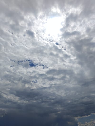 The image displays a cloudy sky, suggesting overcast conditions or a partial break in cloud cover. The sun is partially visible through a gap in the clouds, creating a bright, diffused light source in the upper portion of the frame. The clouds vary in density and color, with some appearing bright white where illuminated by the sun, and others a darker gray, indicating thickness or shadow. Patches of blue sky are visible between some of the clouds, particularly in the middle section of the image. The lower part of the image shows denser, darker clouds, hinting at the possibility of rain or a storm system approaching or passing. The image is a photograph taken from a low angle, looking upwards at the sky, with no visible ground, buildings, or people. It captures a natural atmospheric scene. There is no information within the image itself to indicate its location in Colonița, Moldova.