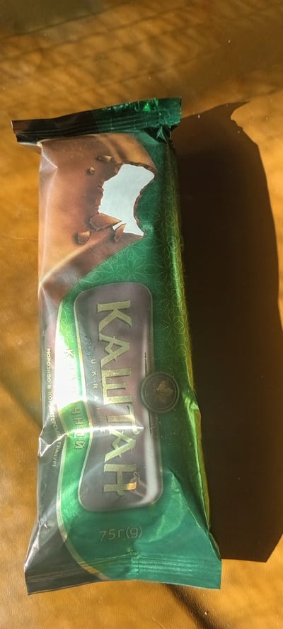 The image shows a close-up, top-down view of a chocolate ice cream bar wrapper on a brown, textured surface, possibly wood, illuminated by a strong light source casting a dark shadow to the right. The wrapper is predominantly green with a pattern of light green geometric shapes. The brand name " КАШТАН " in large, gold, serif letters is visible across the center of the wrapper. Below it, in smaller gold letters, are the words " UKRAINA ". To the left of the main title, in smaller, vertically oriented text, also in gold, are words that appear to be " МОРОЗИВО. В. КОНДИТЕРСЬКИЙ ГЛАЗУРІ " and " ЧУДНИЙ ". The bottom right of the wrapper has the weight indicated as " 75г(g) ". The wrapper has a slightly torn section near the top left, revealing a brown, chocolate-like surface underneath, with some broken pieces visible. There are no people or animals in the image, and the scene is a still life focused on the product packaging. The location in Kovyaghy, Ukraine, is not visually represented in the image itself.