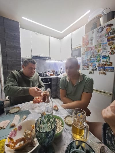 Three men are seated around a table laden with food and beverages. A glass of beer sits beside a green bowl containing peeled nuts. There are plates of various dishes, including fish. One man wears a green shirt, another a gray sweater. A fridge is visible behind them, adorned with decorative magnets. The room is lit by ceiling lights.