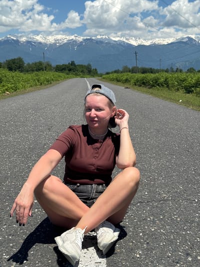 A young woman is pictured sitting on an asphalt road in a scenic outdoor setting, identified as Pakwash, Abkhazia. She is wearing a dark brown short-sleeved collared shirt with buttons, dark shorts, a light grey baseball cap worn backward, and white athletic sneakers. A bracelet is visible on her left wrist, and a ring adorns her left ring finger. Her nails have dark polish.
She is positioned centrally in the frame, sitting with her legs bent and slightly apart, and her arms resting on her thighs. One hand is near her ear. She looks directly at the camera with a neutral expression.
The road stretches into the distance, flanked by lush green vegetation, possibly bushes or low trees, on one side. In the background, majestic mountains rise, some of their peaks distinctly snow-capped under a clear blue sky dotted with scattered white clouds. Utility poles and power lines can be seen in the mid-ground, leading towards the mountainous landscape. The scene is brightly lit by the sun, casting strong shadows from the woman onto the asphalt, indicating a sunny day, likely late morning or afternoon. There is no visible text in the image.