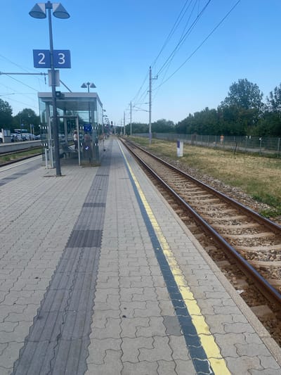 The image depicts a train station platform in Eisenstadt, Austria. The platform is paved with grey interlocking concrete blocks and features a tactile paving strip for the visually impaired. A modern glass-enclosed waiting area with benches is visible on the left side of the platform. Overhead, a blue sign displays the numbers "2" and "3", indicating track or platform numbers. Streetlights are mounted on poles.
Adjacent to the platform are active railway tracks. The tracks are laid on ballast and are bordered by a yellow and black line marking the platform edge. In the background, there is a metal fence separating the tracks from a grassy area and a line of trees. A few cars are parked in the distance on the left. The sky is clear and blue, suggesting fair weather. One person can be seen inside the waiting area, and another person is walking a dog on a leash along a path behind the fence.