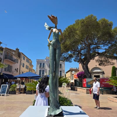 In Saint-Tropez, France, a bronze statue of a slender, fashionably dressed figure stands in a sun-drenched plaza. The statue depicts a woman with her hands on her hips, her attire consisting of a bustier top and flowing, wide-legged trousers, with a scarf trailing from her neck. A woman with long, dark hair wearing a white dress stands facing away from the camera, looking at the statue. To the right, a man in a white polo shirt and navy shorts is looking towards the statue. In the background, a large pine tree with dense foliage dominates the scene, with buildings typical of a Mediterranean town lining the plaza. Blue umbrellas from outdoor cafes and tables with chairs are visible, suggesting a vibrant street-side dining area. A red fire hydrant stands near the man. Signage for an exhibition titled "HENRI PERSON, une personnalité tropézienne" is displayed on red panels. The sky is a clear, bright blue, and a seagull is visible in the upper left corner.