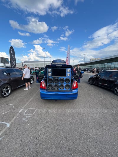 An outdoor, daytime scene is depicted, featuring a car show in Nizhny Novgorod, Russia. The primary focus is a blue sedan with its trunk open, revealing a complex audio system installed within. This system includes multiple speakers, some of which are labeled "AURA," arranged in a custom enclosure. Above the speakers, a large screen is visible, and a blue light strip illuminates the setup.
Several people are present, some standing near the vehicles, observing or interacting. One man in a white t-shirt and black shorts stands next to a dark-colored sedan to the left. Other individuals are seen in the background, near other cars and structures that suggest a large event venue, possibly a stadium or exhibition center, which is characteristic of the Nizhny Novgorod area.
The environment is an asphalt lot, with parking lines and markings visible on the ground. Two large flags are planted to the left and right of the blue car, one black with white lettering and the other white with red lettering. The sky above is blue with scattered white clouds, indicating fair weather. Other cars, including a black sedan to the right, are parked in the vicinity, suggesting a gathering of vehicles. The overall impression is that of a car enthusiast event focused on modified sound systems.