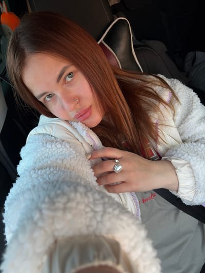 A young woman is depicted in a close-up, upward-angled selfie, positioned inside what appears to be a car. She has long, reddish-brown hair and green eyes, with a septum piercing (nose ring). Her lips are slightly parted in a neutral expression. She is wearing a white, fluffy, sherpa-style jacket with a light-colored, possibly lilac, zipper lining, over what seems to be a lighter-toned underlayer. A prominent silver ring with an irregular, pearl-like or organic-shaped white stone is visible on her left ring finger. Her fingernails are painted a light, neutral color.
The setting is an interior, characterized by dark car seats with white piping and a visible black seatbelt draped across her body. The background is dark and blurred, suggesting the confined space of a vehicle. A light-colored fabric with faint red text, possibly reading "juste," is partially visible behind her arm. An orange, plush object can be glimpsed in the lower left background, near what looks like a headrest. The lighting suggests daytime, but the source is diffuse, possibly from inside a car or an overcast sky. The overall activity is a self-portrait or a casual photo taken while seated in a car, likely in Alexandrovka, Russia.