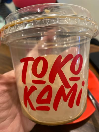 A clear plastic beverage cup with a transparent domed lid is held by a human hand, primarily visible on the left side of the frame. The cup features the brand name "TOKO KAMI" in prominent red block lettering. A small amount of light brown liquid residue, indicative of a previously consumed beverage, remains at the bottom of the cup and is present on the interior of the lid. The background is out of focus, revealing a dark brown surface, likely a table. To the right of the cup, a bright orange or red circular plate is partially visible, with a metallic utensil, possibly a spoon, resting upon it. Further blurred elements in the background suggest an indoor commercial establishment, such as a cafe, located in Pontianak, Indonesia.