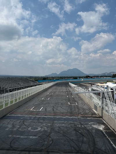 The image depicts a section of the Autódromo Miguel E. Abed race track in Amozoc de Mota, Mexico. The track surface is asphalt, with visible tire marks and painted lane markings. To the right of the frame, a concrete barrier with faded lettering is present. On the left side, stadium seating and metal structures can be seen. In the background, a mountain range is visible under a sky with scattered clouds. The image is rotated 90 degrees clockwise.