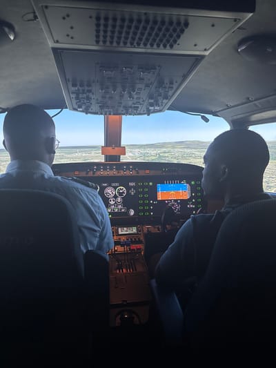 The image displays the interior of a flight simulator cockpit, implicitly located in Port Alfred, South Africa. Two male individuals are seated in the pilot and co-pilot positions, observed from a rear perspective. The individual in the left seat wears a light-colored pilot uniform shirt with visible epaulets and eyeglasses, looking forward at the simulated environment. The individual in the right seat wears a dark long-sleeved shirt, also focused forward.

Above the occupants, an overhead panel is visible, containing rows of numerous switches and buttons. The instrument panel directly ahead of the occupants features multiple illuminated digital displays presenting flight data, navigation information, and engine parameters, flanked by several analog gauges. A central pedestal between the two seats contains throttle levers, trim wheels, and additional control inputs.

Through the cockpit windshield, a simulated external landscape is depicted. A runway with distinct markings is visible straight ahead, surrounded by a mixture of green and arid brown terrain featuring scattered small structures. The sky above is clear and blue, with minimal cloud cover. The scene indicates a flight training or simulation exercise in progress.