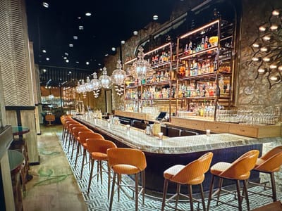 The image depicts an interior view of a dimly lit bar with a long, curved counter. The counter is made of polished stone and has a dark blue, ribbed base. There are several orange bar stools with wooden legs arranged along the counter. Behind the counter, shelves are stocked with a variety of bottles, primarily spirits and liqueurs, illuminated by overhead lighting. Crystal chandeliers hang from the ceiling, adding to the ambiance. The flooring consists of a patterned tile design. The overall impression is that of a sophisticated establishment. The location in Ellwangen, Germany, is not specified within the image itself.