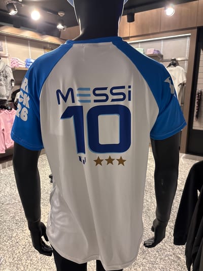 A black mannequin wearing a white t-shirt with blue sleeves is displayed in what appears to be a sports merchandise store. The t-shirt has the name "MESSI" printed in large blue letters across the back, with the number "10" prominently displayed below it. Three gold stars are also visible below the number. In the background, shelves are stocked with various clothing items, including pink hoodies and grey hoodies. The store has a modern interior with suspended lighting fixtures. The floor is a speckled grey. The t-shirt itself is white with blue raglan sleeves, and the graphics are bold and clear. The lighting is bright, illuminating the merchandise effectively. The overall impression is that of a retail environment focused on sports apparel. This scene is observed inside a shop within the Aeropuerto Internacional Ezeiza (EZE) in Argentina, a busy international airport.