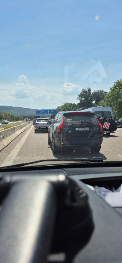 The image shows a view from inside a vehicle on a highway, likely in Germany, due to the signage and vehicle license plates. The highway is congested with several cars, indicating a traffic jam. The sky is blue with scattered clouds. Trees and hills are visible in the background. One of the vehicles in the foreground is a dark-colored Volvo XC90 with a license plate that reads "EL6 58CC". Another car in front of it is a silver Volkswagen with a license plate "KA-NA 9250". A white van is also present. The overall scene depicts a typical highway traffic situation on a sunny day. The image does not provide specific details that would confirm the location as Karlsruhe, Germany, although the presence of German license plates suggests a German setting.