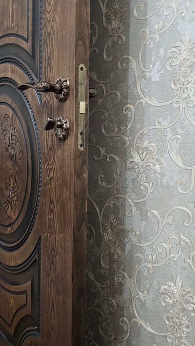 The image shows a partially opened wooden door with ornate bronze handles and a metallic lock mechanism. The door is made of dark, textured wood with carved patterns. To the right of the door is a wall covered in ornate wallpaper with a damask pattern in shades of grey and beige. The scene appears to be indoors, possibly in a residential hallway or entrance, with no visible signs of the city of Yandare, Russia. There are no people or actions depicted in the image.