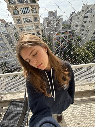 A high-angle portrait depicts a young female with light brown hair, eyes closed, and her head tilted slightly downward. She wears a dark blue hooded sweatshirt with white drawstrings and a faint graphic on the chest. Freckles are visible on her nose and cheeks. A small stud earring is present in her right ear. Her lower body shows dark pants and brown boots.

The subject stands on a balcony or rooftop terrace, indicated by a prominent white safety net grid that covers much of the foreground and midground. The balcony floor features a patterned surface, possibly wooden or tiled, with distinct shadows cast by sunlight. A dark wicker chair is partially visible on the left.

The background reveals a dense urban environment under an overcast sky. Numerous multi-story buildings populate the cityscape. A distinctive, centrally located building features a facade with horizontal bands of colorful panels in red, orange, green, blue, and purple. This building is identifiable as Condomínio Residencial Itapema (Edifício Itapema) at Rua Mariante, 396, Moinhos de Vento, Porto Alegre, Brazil. To the left, a tall, cream-colored building with multiple balconies is prominent. Other grey high-rise structures and scattered green trees are visible throughout the urban panorama, consistent with the Moinhos de Vento or Bela Vista neighborhoods.