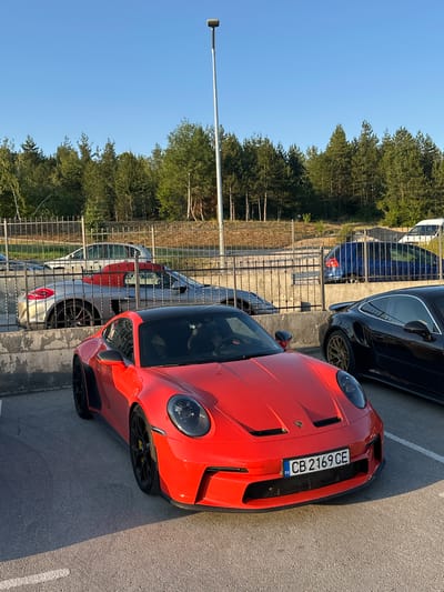 The image shows a parking lot with several cars and trees in the background. The main subject is a bright orange sports car parked in the foreground, angled towards the right side of the frame. It has a black roof and black accents on the body. A license plate is visible on the front of the car, which reads "CB 2169 CE BG". To the left of the orange car, a silver car is partially visible. Behind the silver car, a black sedan is parked, and further behind that, a dark blue car is visible. The cars are parked in a lot paved with asphalt. A metal fence runs along the left side of the parking lot. Beyond the fence, a dense line of green trees forms the background, with a clear blue sky above. A street lamp is visible on the far left side of the image. The lighting suggests it is daytime, likely late afternoon or early morning due to the angle of the shadows. The scene appears to be in Kerenite, Bulgaria, as indicated by the license plate and the provided location context. There are no people visible in the image, and the cars appear to be parked and stationary.
