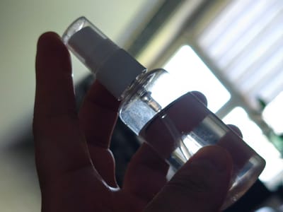 A close-up, silhouette-style shot shows a person's hand holding a small, clear spray bottle. The bottle has a white nozzle and cap. Light streams in from the background, creating a bright, out-of-focus window with vertical blinds. The overall lighting suggests an indoor environment with bright natural light from a window. There is no information in the image that allows for a determination of the location within Зажэвіцкі сельскі Савет, Belarus.