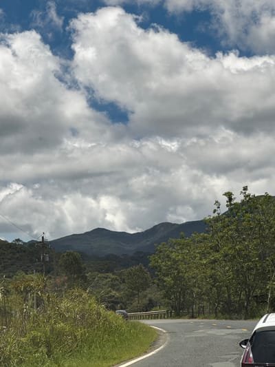 The image shows a winding road in a mountainous, rural area. Lush green vegetation covers the landscape, with tall grasses and trees lining the road. In the distance, dark green mountains rise under a dramatic sky filled with white and gray clouds. A utility pole with electrical equipment stands on the left side of the road, and a dark-colored car is visible in the distance on the right, driving away from the viewer. The road itself is asphalt and appears to be in good condition, with yellow reflectors marking its edge. The overall impression is one of natural beauty and remote tranquility. This scene is located in Đưng K'Si, Vietnam.
