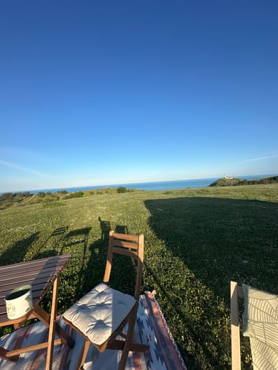 The image shows a picnic scene on a grassy field overlooking the sea. A clear blue sky is visible above the horizon. Two wooden chairs are set up on a blanket. A mug is on one chair. The landscape includes a grassy hill leading to the sea. A building is visible in the distance. The city is Emona, Bulgaria. There are no visible people in the image.