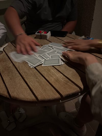 The image captures a casual card game taking place in dim lighting, likely during the evening or night. A round wooden table with visible grain and slats serves as the central surface for the game.
At least two people are visible, actively engaged with the cards. On the left, a person wearing a dark grey short-sleeved shirt has their arm and hand reaching towards a spread of playing cards. A silver bracelet adorns their wrist, and a white watch with a gold face is also visible. Next to this person, a dark-colored smartphone lies on the table, and a stack of UNO cards, bound by a rubber band, sits nearby. The text "UNO" is clearly visible on the side of this deck.
Another person's hands are seen from the top-right, also interacting with the scattered playing cards. This individual appears to be wearing light-colored clothing and white sneakers, partially visible under the table. Another pair of feet, wearing light-colored sandals with a decorative element, are visible on tiled flooring in the bottom-right corner.
Numerous playing cards, showing their blue-patterned backs, are fanned out and scattered across the wooden table. The hands of both individuals are blurred, indicating motion from shuffling, dealing, or playing. A small blue object with the partial text "DECK" is barely visible near the top-right hands. The background is dark and indistinct, while the visible flooring under the table suggests an indoor or sheltered outdoor setting. The scene conveys a relaxed, social gathering centered around card games.