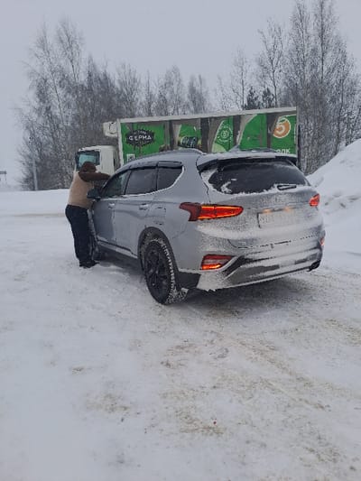 A gray car is parked on a snowy road. Snow covers parts of the car, especially the rear. A person is visible near the driver's side door, possibly brushing snow off the car. A large green truck with lettering in Cyrillic is parked behind the car, near bare trees. The background sky is overcast.