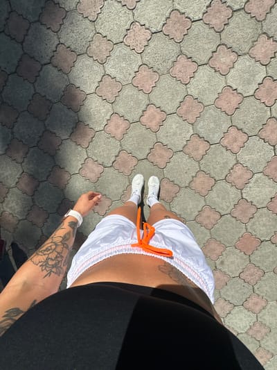 An overhead shot shows a person's legs and torso standing on a patterned tile surface in the city of Duvankul', Russia. The tiles are a light grey with decorative pink clover-like shapes. The person is wearing white athletic shoes, white shorts with an orange drawstring, and a black top. A tattoo is visible on the person's left arm and a white smartwatch is on their left wrist. The ground is partially covered in shadow from an unseen object.
