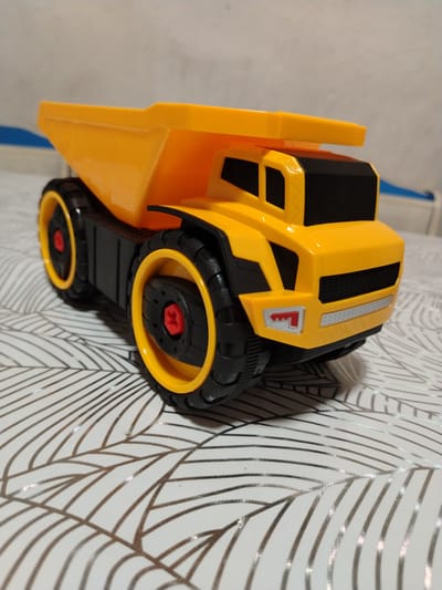 A yellow toy dump truck is positioned on a white surface with a pattern of thin, parallel lines resembling leaves or veins. The truck is viewed from a three-quarter angle, with its front facing towards the right of the frame. The dump bed is raised, and the truck has large black tires with yellow rims. The cabin of the truck is black and yellow. The background is out of focus, showing a light-colored wall. There is no indication of the city of Жернов ауылдық округі, Kazakhstan in the image.