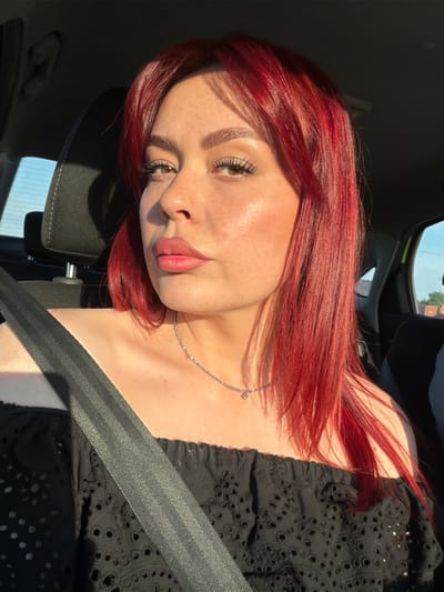 In the city of Узюково, Russia, a woman with vibrant red hair is seen inside a vehicle. She is wearing a black, off-the-shoulder top with intricate eyelet detailing and a silver necklace. A gray seatbelt crosses diagonally over her shoulder and chest. The natural sunlight casts a warm glow on her face, highlighting her freckles and makeup. The background shows the interior of the car, including a headrest and a glimpse of the window, suggesting a daytime journey.