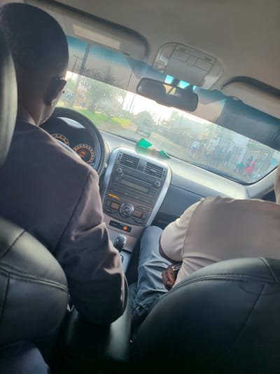 The image shows the interior of a car, likely in Karu, Nigeria, as suggested by the presence of palm trees and buildings common in the region. Two men are visible. The driver, wearing a dark suit jacket, is in the foreground on the left, with his back to the camera. He is looking forward, suggesting he is driving. His hands are on the steering wheel. The passenger, wearing a light-colored t-shirt and jeans, is in the passenger seat on the right, also with his back to the camera. He appears to be looking out the windshield.
The car's dashboard is visible, with the steering wheel, instrument cluster, and radio. Outside the car, through the windshield, a street scene is depicted with several people and motorcycles. The lighting suggests it is daytime, possibly late afternoon due to the warm sunlight. The overall impression is a candid shot of people traveling in a car in a semi-urban or rural setting in Nigeria.